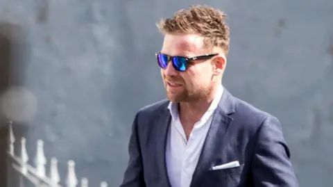 Mark Pearce Rory Trainor, pictured in 2015.  He has short, blonde hair and dark mirrored sunglasses.  He is wearing a navy suit jacket and a white open nect shirt. 