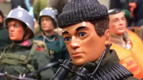 BBC The head and shoulders of an Action Man figure, wearing a black beanie hat and binoculars round his neck. Several other military figures can be seen behind out of focus.