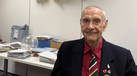 An elderly man with short white hair, large wire-framed glasses and a fairly narrow face smiles at the camera. He wears a red shirt, red, black, white and yellow tie, and black blazer with various poppy and remembrance badges. Behind him on a table are many stacks of cards in envelopes.