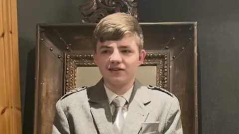 Police Scotland a teenage mail with short fair hair is wearing a grey formal jacket with white shirt and grey tie