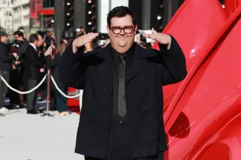 Getty Images Caleb Hearon wears a black jacket, shirt, tie and trousers while gesturing with his hands on the red carpet.