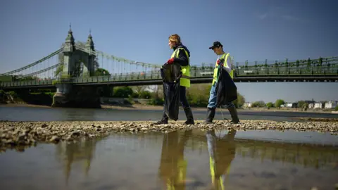 Two volunteers can be seen participating in a clean-up of a Thames riverbank. They are stood on a beach under a green bridge wearing yellow hi-vis vests putting littler into black bin bags using litter pickers.
