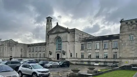 A view across a car park in front of Southampton City Council's main building. It is a large greystone building with a central section, nearby clock and two wings with Georgian windows extending outwards either side. 