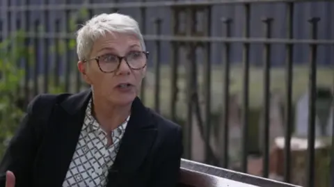 Alison Hinds wearing a dark jacked and white chequed blouse with dark glasses, sitting in front of a fence