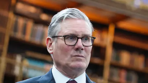 Keir Stsramer inside the British Museum looking serious wearing a suit and tie, head and top of shoulders shown. 