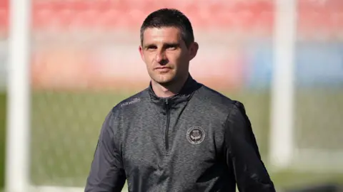 PA Media A man with short dark hair, wearing a grey sports top, on a football pitch. An out of focus goal is visible behind him.