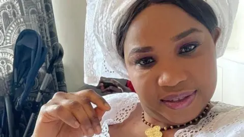 Ndata Bobb is sitting in a room in a house. She is looking into the camera and is smiling. She has a hand raised close to her face. She is wearing a white outfit decorated with embroidered circular patterns.