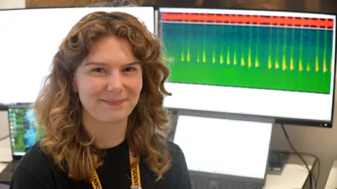 Jamie Niblock/BBC Grace Tinklin is smiling at the camera. She has wavy, shoulder-length brown hair and is wearing a dark jumper and a lanyard around her neck. There is a computer screen in the background with coloured lines and patterns on it.