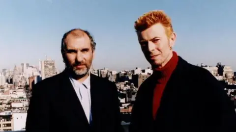  Bowie At Fifty - Picture shows Alan Yentob (left) and David Bowie. Yentob. The pair, both wearing black suits, are pictured with the London skyline behind them.