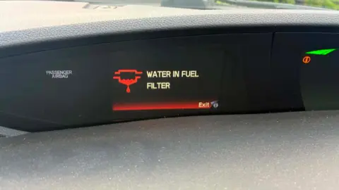 David Bush It shows the dashboard of a Honda car. On this, it says 'Water in fuel filter' in white block capital. There is a red trim and a red engine emblem with a red water drop below it. 