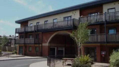 The courtyard entrance to an apartment complex, with balconies seen running outside windows over the two-storey building. Trees can be seen outside in a gated seating area. 