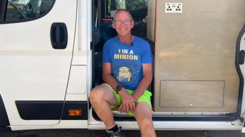 BBC Mark Ferbrache pictured wearing a blue Minions-themed T-shirt, bright green shorts and hiking shoes sitting on the open side doorway of a white camper van. The van’s interior is visible, showing wood-panelled storage units, a power outlet and a sleeping area toward the back.