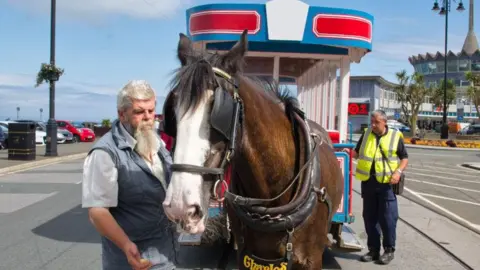 MANX SCENES A tram driver with a grey beard and hair standing next to the horse with the tram and the Sea Terminal building behind.