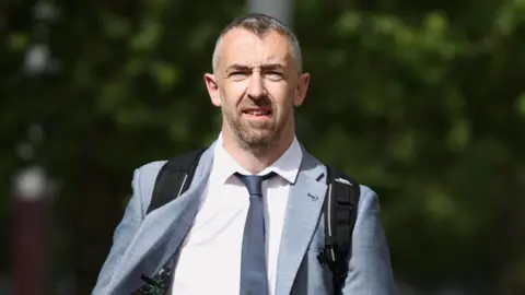 PA Media A man with greying short hair and a greying short beard. He is wearing a light grey suit with a darker grey tie. He is carrying a black backpack. Behind him are some trees.