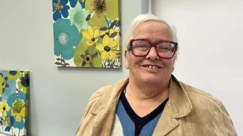 Charlotte Vowden/BBC Diabetic Sandra McDermott, who was diagnosed with the disease 20 years ago, is stood in front of floral artworks that are hung on the wall. She has short hair and is wearing a cream jacket, blue and black jumper. Her glasses have a tartan pattern on their frame.