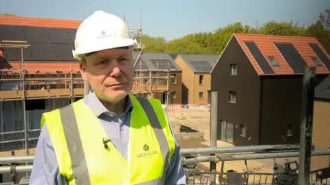 Ian Pritchett is standing on scaffolding at a building site. He is wearing a white hard hat and a yellow hi-viz vest over a grey shirt. The homes in the background are partially completed and all have solar panels on the roof.
