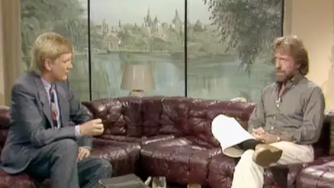 A man on the left wears a suit sitting on a couch facing a man on the right wearing a light brown shirt and light pants.