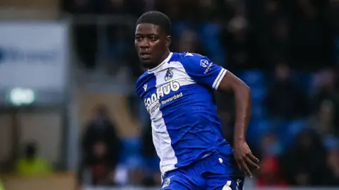 Clinton Mola on the pitch. He wears the blue and white Bristol Rovers strip. Fans can be seen in the stadium behind. 
