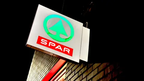 Getty Images a Spar supermarket outside store sign. It's night and the sign is lit up. The logo is a green tree in a circle.