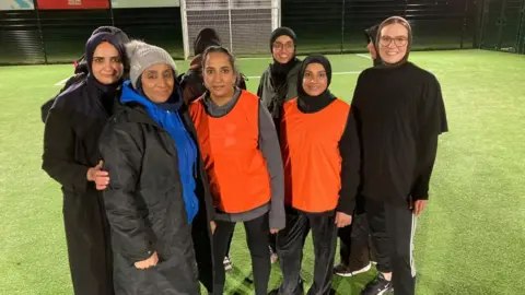 A group of women are looking active. Two people are wearing orange shirts and seem to be in the same team. There is a woman in a blue jumper to the left. Everyone is smiling.