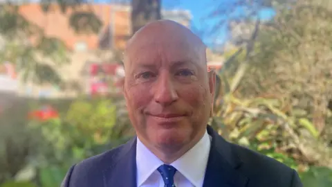 A man smiling at the camera. He is wearing a navy suit jacket with a white shirt and a blue tie with white polka-dots. He is bald, and is standing outside in front of a number of trees and bushes.