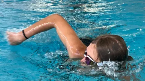 A swimmer wearing goggles performs freestyle in an indoor pool, with one arm raised during the stroke and water splashing around the head. Blue water and red, white and blue lane dividers run alongside the swimmer in a marked swimming lane.