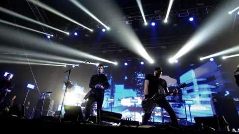 Pendulum on stage- Two men dressed in black with guitars with a blue city skyline background