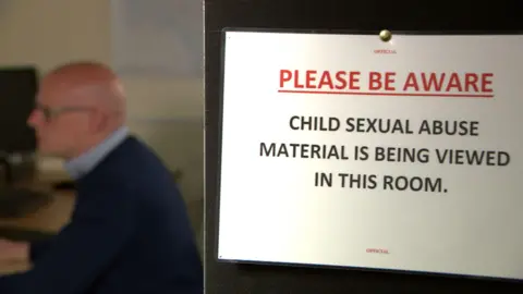 David Murray sits in the distance working at his desk as we look into the office from the doorway. The open door has a large sign pinned on it which says "Please be aware. Child sexual abuse material is being viewed in this room".