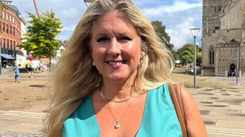 Alison Sheridan wearing a turquoise dress and silver jewellery with a tan handbag strap over her shoulder and standing in front of Exeter Cathedral.