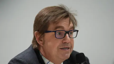 PA Nik Johnson is looking to the side of the camera while speaking into a microphone. He wears square-framed black glasses and a suit. He has a moustache and brown hair. 