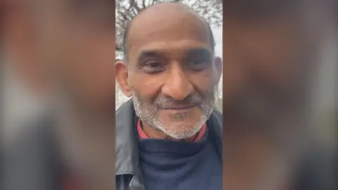 A man with a short grey beard is smiling. He is wearing a shirt with a red collar, a blue jumper and a black jacket.