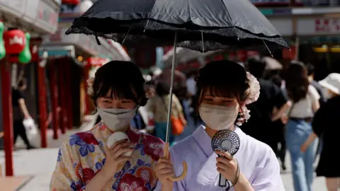 Two women, dressed in summer kimonos, hold an umbrella and portable fans as they walk through a Tokyo street.