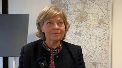 Warwickshire County Council leader Izzy Seccombe in a dark suit jacket standing in an office, with a map behind