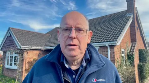 Donald Potter wearing a blue fleece jacket stands in front of a red brick bungalow. He has a balding head with white hair and is wearing silver-rimmed glasses.