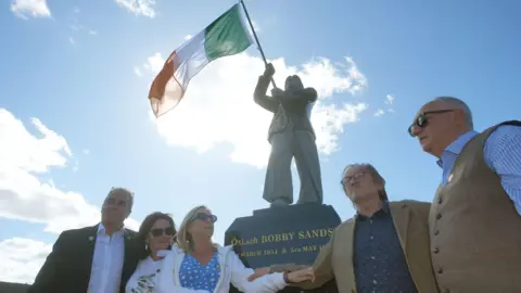 Brian Lawless/PA Three men and a woman stand should to shoulder in front of a statue.