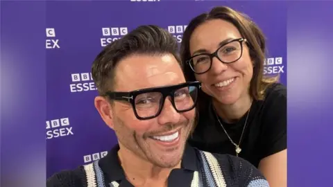 A man with a brown quiff and thick-rimmed black glasses smiles at the camera. A woman with dark hair who is also wearing black-rimmed glasses stands behind him and is also smiling.