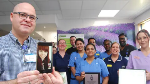 Kettering General Hospital Group of medical people in a hospital ward. A man with very short hair and glasses, wearing a blue and white shirt and a stethoscope is holding a smartphone on which Kirstie, with long red hair, is visible. Beyond him are various nurses and healthcare assistants wearing mainly blue uniforms.