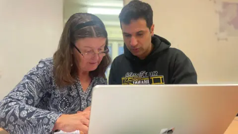 Simon Thake A young man with dark hair and an elderly woman sit down in front of a laptop