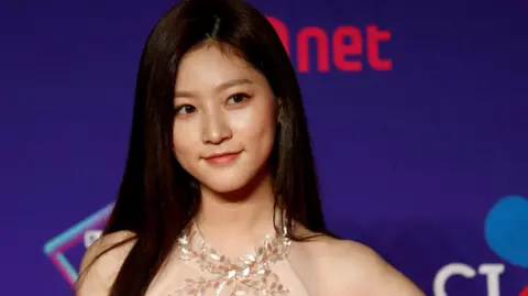 Kim Sae-ron, a young woman with long dark brown hair and brown eyes, stands in front of a purple wall on a red carpet, wearing a pink sleeveless dress.