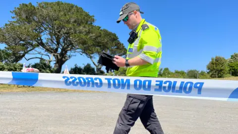 BBC Image is taken behind blue and white police tape. In front of the tape a police officer wearing a high viz top and he is holding an electronic tablet.
