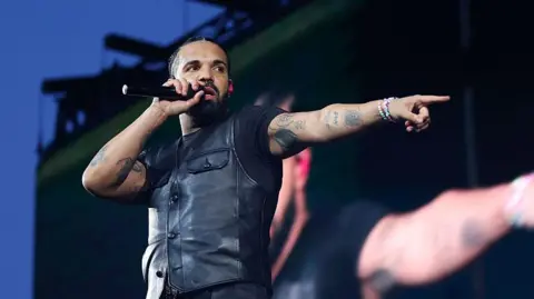 Drake performs on stage, holding a microphone to his mouth. He's pointing at a fan in the crowd with his free hand. He wears a black leather vest over a black t-shirt.