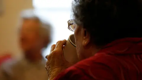 The left side of an elderly lady's face as she drinks from a mug. There is a ring with a large gem on her left hand and she is wearing glasses. She has dark grey hair. Sitting opposite her, but blurred, is another lady who has pale grey hair and is wearing glasses.