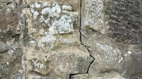 Alan Richardson A crack running down stonework in a jagged line. The stones are weathered with lichen on them.