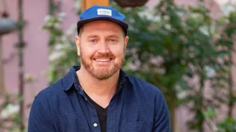 Helpu A man with a ginger beard wearing a blue baseball cap, black t shirt and blue shirt and smiling at the camera. He is sat in front of some green plants.
