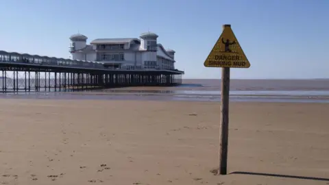 Getty Images A triangular yellow warning sign on top of a signpost on Weston-super-Mare's beach, next to the pier. The signs says 'danger, sinking mud' with a silhouette image of a man up to his knees in the sand.