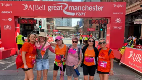 The Great Run Company Angela (middle) stands with five members of her running team in front of the AJ Bell Great Manchester Run finish line. All but one wear orange T-shirts with Maggie's written on the front in white letters