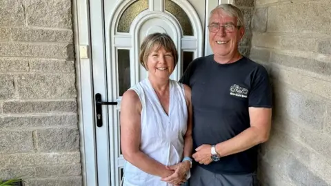 A man and a woman are standing next to each other in front of a white door to their home. They are both smiling.