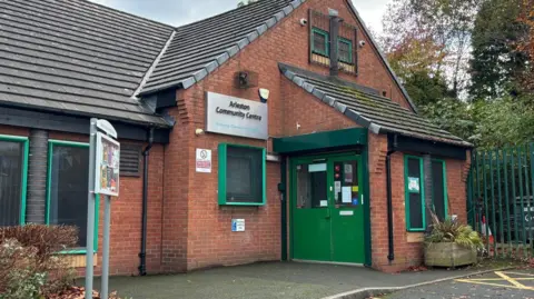 A brick-built community centre with green window frames and doors and a silver sign on the front with "Arleston Community Centre" in black lettering.