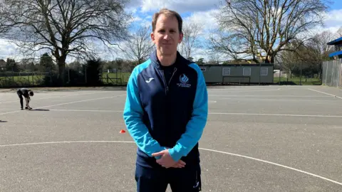 Leanne Rinne/BBC PE teacher Nathan Sasson. Nathan is stood with his left hand over his right, and is wearing a school branded navy and pale blue tracksuit. He is smiling to camera and is stood in the school playground.