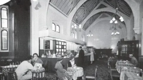 John Huggon/Network Rail A black and white image from about the 1960s of the former first class waiting room at Carlisle railway station. A number of people are sat at tables covered by gingham cloths. Two people are standing at the bar on the left. The building has vaulted ceilings painted in white, with some of the wooden structure visible. Large pendant lights hang from the high ceiling.